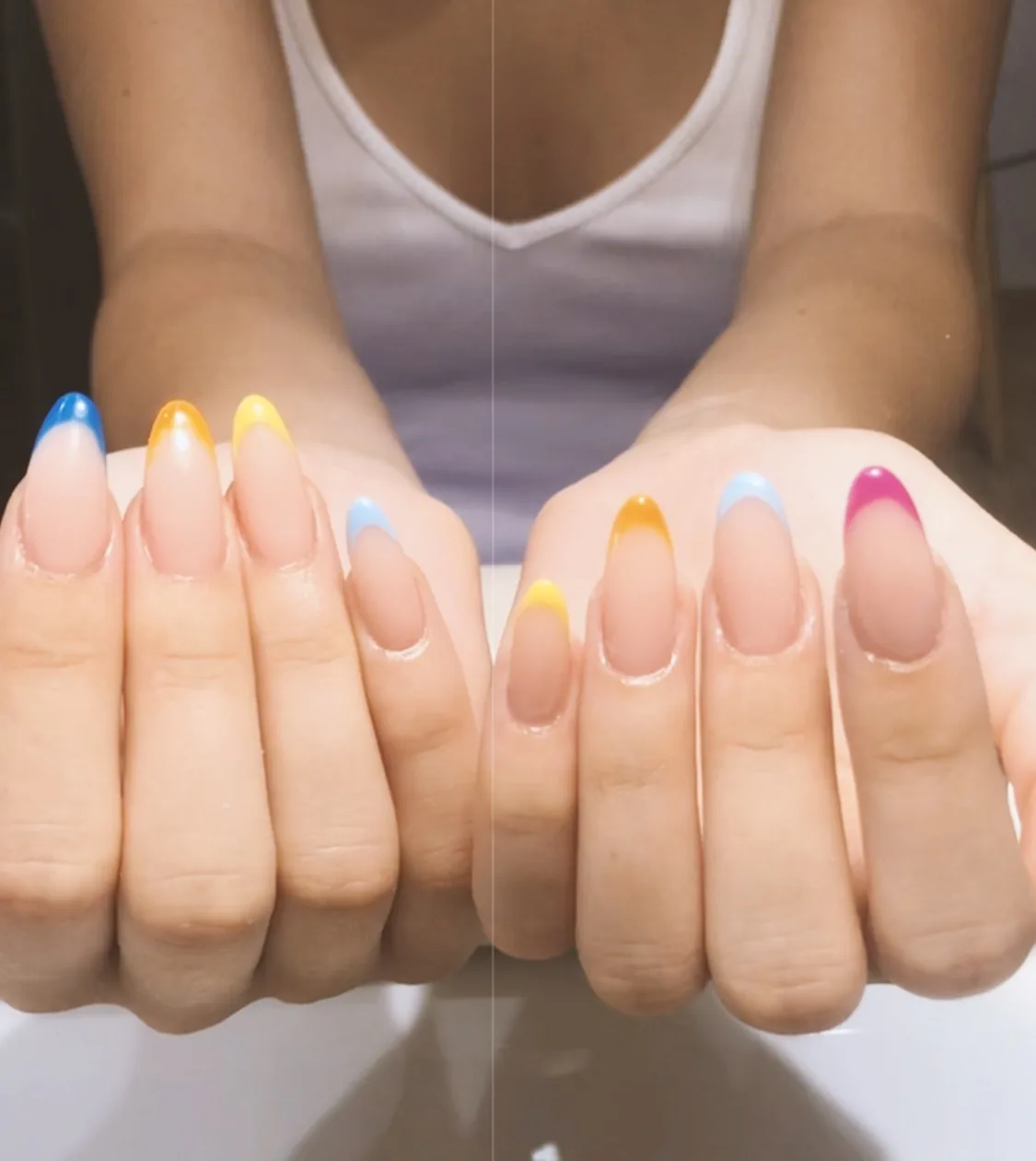 Deux mains posées au-dessus de la table du salon, ongles amande en french tips pastel bleu, jaune et rose.