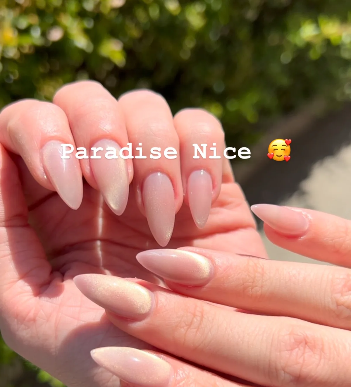Main aux ongles amande nude nacré, photographiée en extérieur devant une haie.